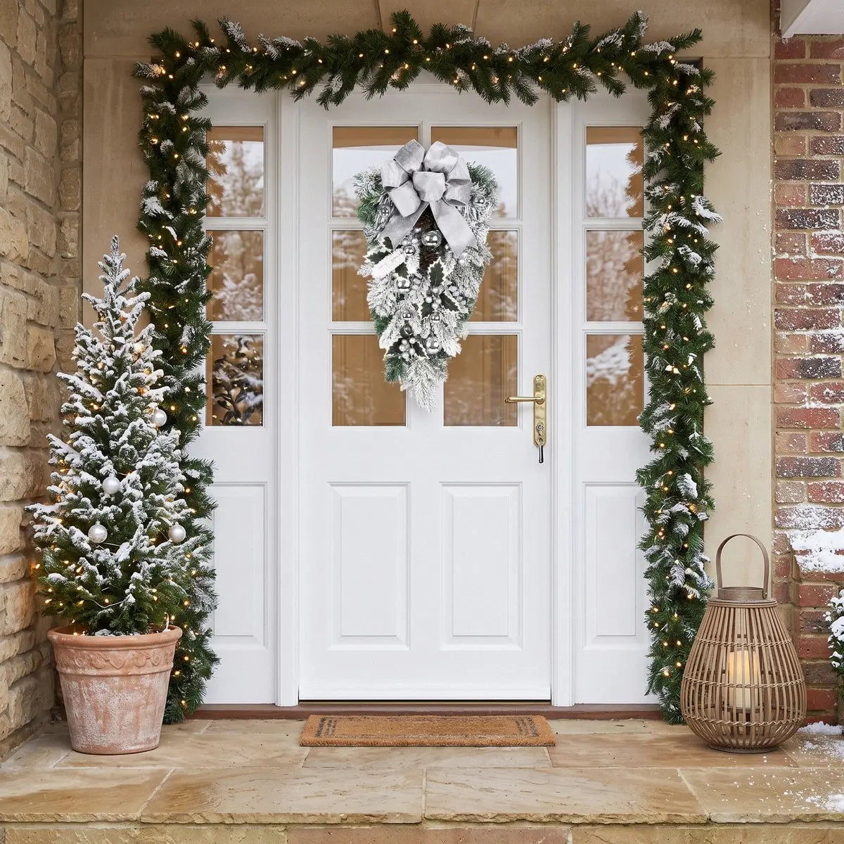 White Christmas teardrop swag with silver bow, silver ball ornaments, frosted pine branches and white berry clusters, hung on a white front door framed by lit pine garland, with a snow-dusted potted Christmas tree and wicker lantern on the porch