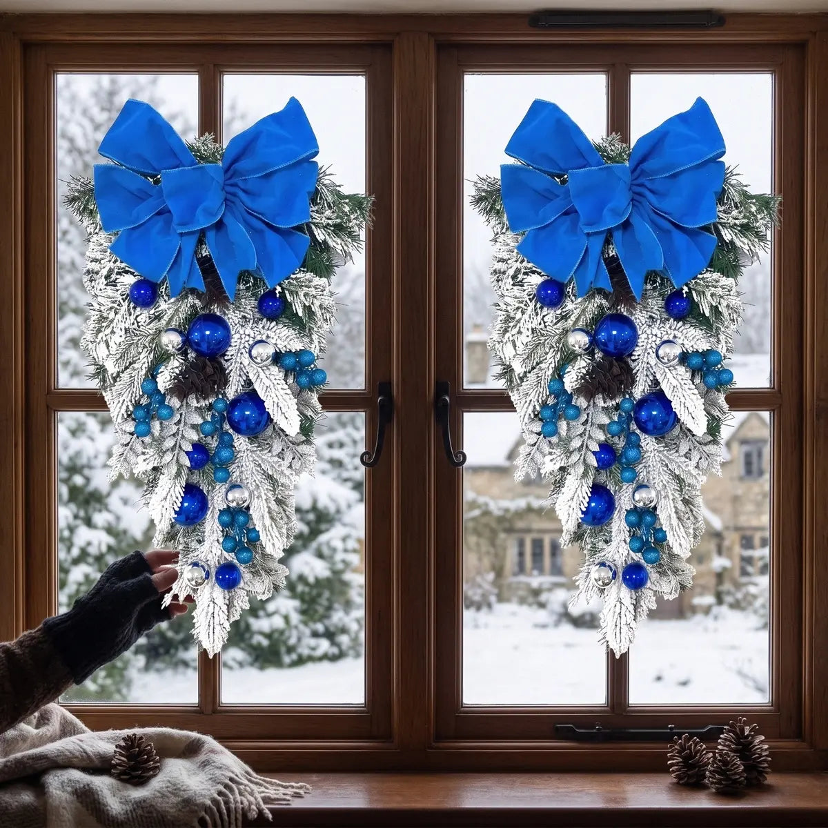 Two blue Christmas teardrop swags with royal blue velvet bows, blue and silver ball ornaments and pine cones, hung on a dark wood double window overlooking a snowy winter landscape with a stone manor house, with pine cones and a knit throw on the windowsill
