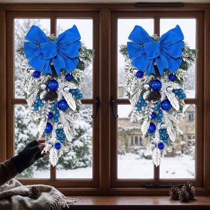 Two blue Christmas teardrop swags with royal blue velvet bows, blue and silver ball ornaments and pine cones, hung on a dark wood double window overlooking a snowy winter landscape with a stone manor house, with pine cones and a knit throw on the windowsill

