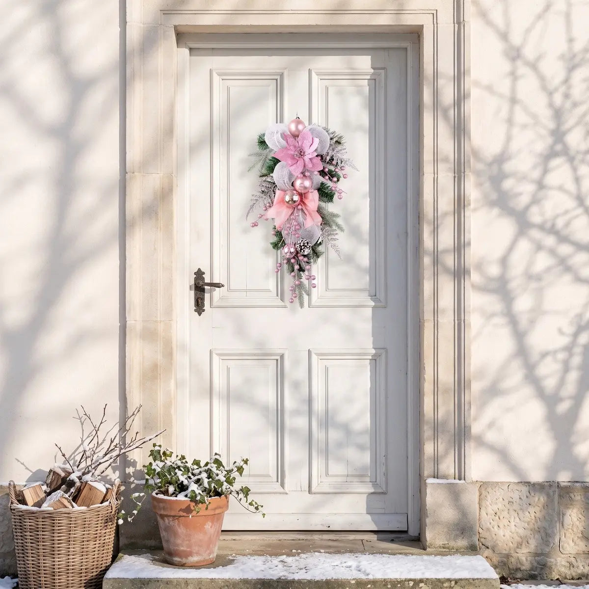 Pink artificial Christmas teardrop swag with poinsettia, mesh bow and ball ornaments, hung on a white front door with winter tree shadows and potted plants
