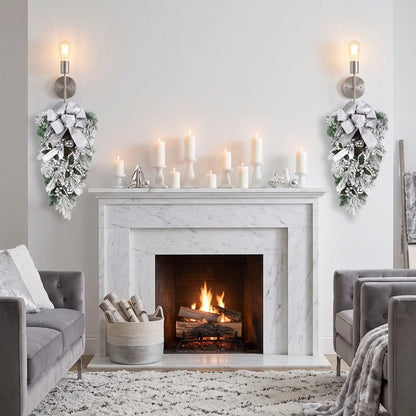 Pair of white Christmas teardrop swags with silver bows and silver ball ornaments, hung symmetrically on either side of a marble fireplace with white pillar candles on the mantel, in a modern silver and white Christmas living room
