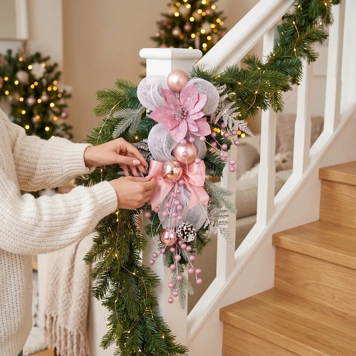 Pink Christmas teardrop swag with poinsettia, mesh bow and rose gold ball ornaments, displayed on white stair railing newel post with garland and lit Christmas tree in background
