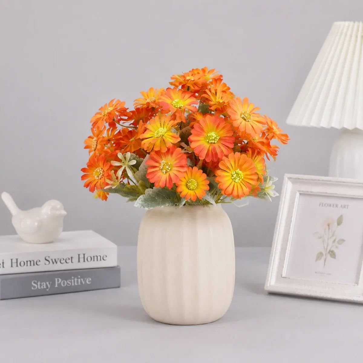 Orange gerbera daisy artificial flowers bouquet arranged in a white ceramic vase on a desk with books and home decor