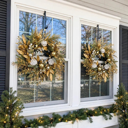 Two 19.7 inch gold Christmas wreaths with gilded golden leaves, gold and silver ball ornaments, white frosted pinecones and silver berries, hung symmetrically on two white-framed exterior windows with black shutters, above a lit pine window box garland, on a grey farmhouse exterior in winter