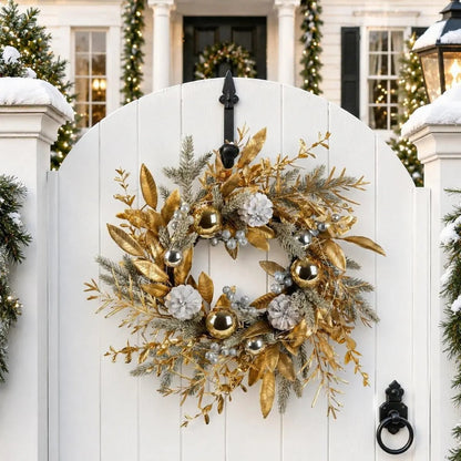 19.7 inch gold Christmas wreath with gilded golden leaves, shiny gold and silver ball ornaments, white frosted pinecones and silver berry clusters, hung on a white arched garden gate with a black iron hook, with a snow-covered colonial-style home decorated with lit pine garlands and Christmas wreaths in the background