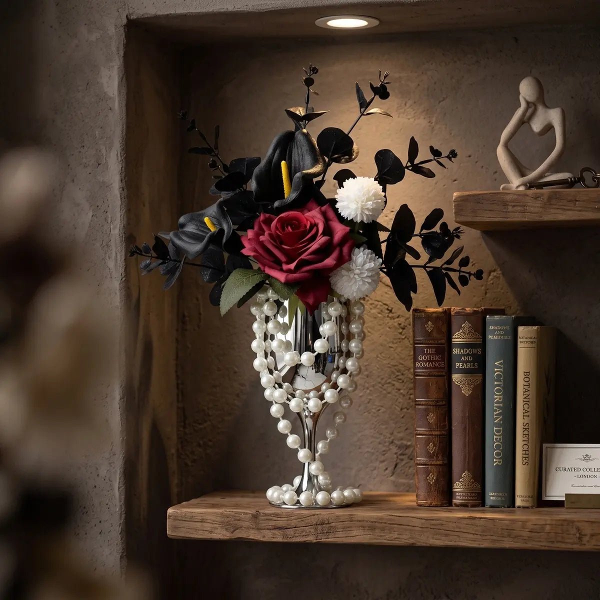 Red rose gothic artificial flower arrangement with black calla lilies in pearl-draped silver goblet vase on a dark styled surface