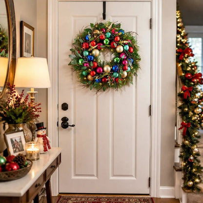 17.7 inch lighted Christmas wreath with multicolor red green blue gold pink and teal ball ornaments, gold cone pendants, gold snowflake picks and eucalyptus leaves with warm LED lights, hung on a white interior front door with a black over-door hook, beside a dark wood console table styled with a snowman figurine, a gold lamp, a glass hurricane candle holder, a pinecone bowl and red Christmas ball ornaments, with a lit staircase garland with red bows visible in the background
