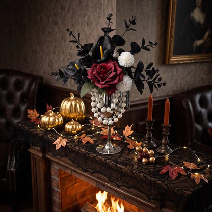Orange rose gothic artificial flower arrangement with black calla lilies in silver goblet vase styled beside a lit fireplace with Halloween decor