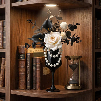 Gothic faux flower arrangement with white rose and black calla lilies in pearl-draped silver goblet vase displayed on a dark wood bookshelf