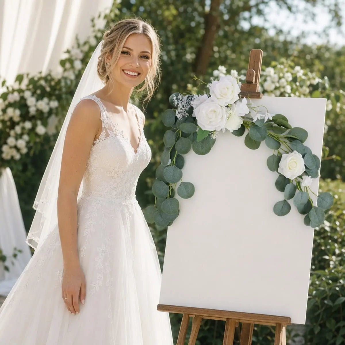 Smiling bride in white lace gown standing beside welcome sign board decorated with white rose silver dollar eucalyptus swag set in outdoor garden wedding – IPOPU